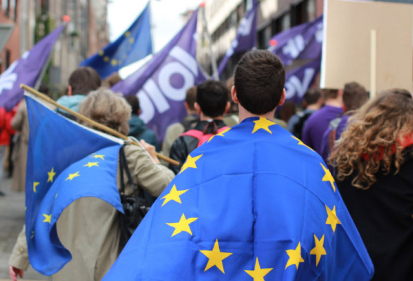 A man in the foreground wearing a European Flag. More European flags are seen in the background with people also waving Volt flags.