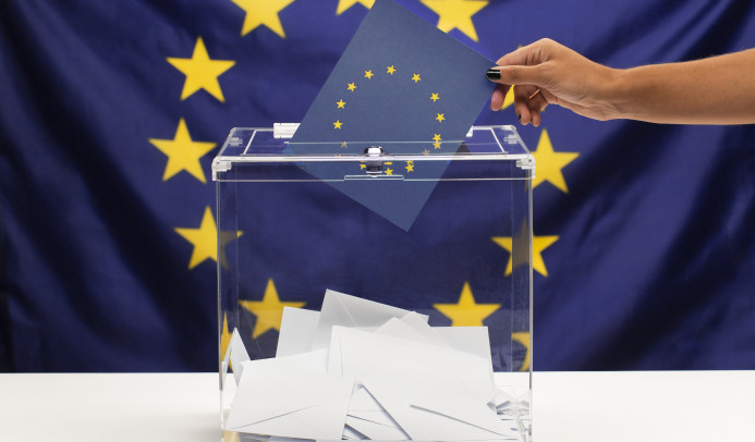A hand putting a voting ballot in an urn, the european union flag in the background